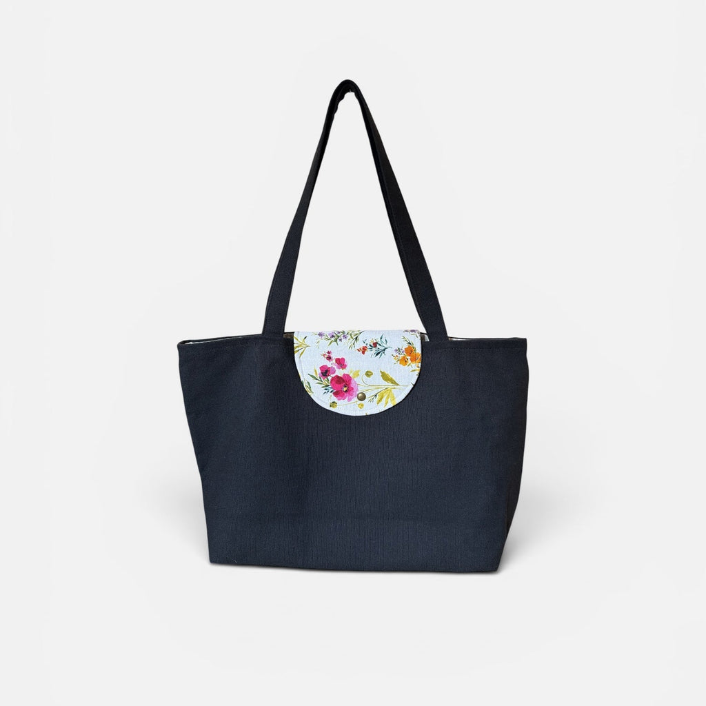 A black canvas tote bag with black shoulder straps and a circular floral patterned detail on the front, set against a plain white background.