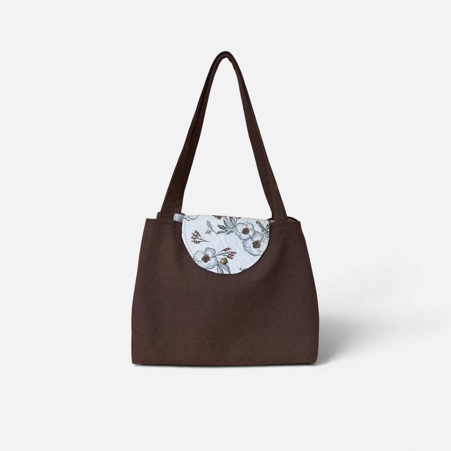 A dark brown tote bag with a circular white and gray floral patterned accent on the front, featuring dual shoulder straps, set against a plain light gray background.