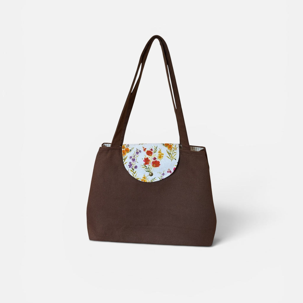A dark brown tote bag with a floral-patterned circular flap detail on the front, featuring dual shoulder straps, set against a plain light gray background.
