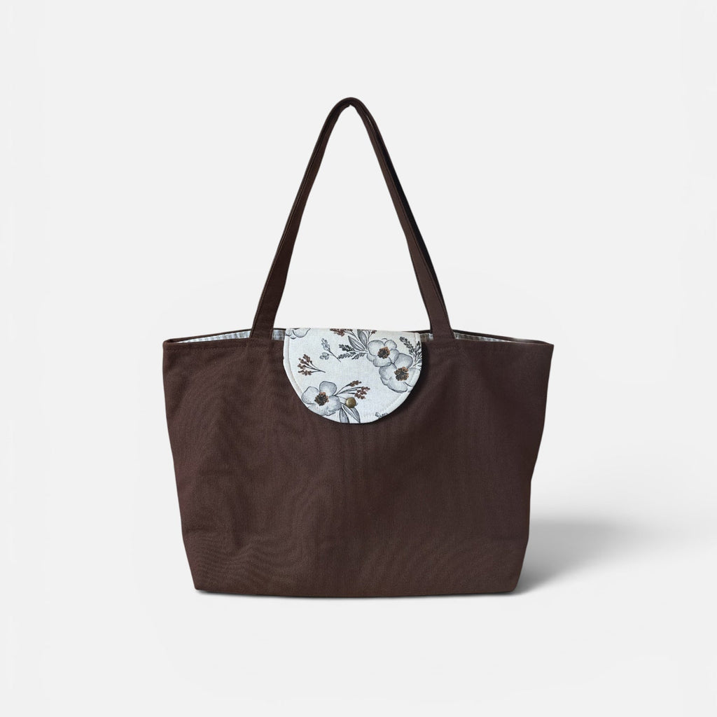 A dark brown tote bag with a circular white floral patch on the front and two shoulder straps, photographed against a clean white background.
