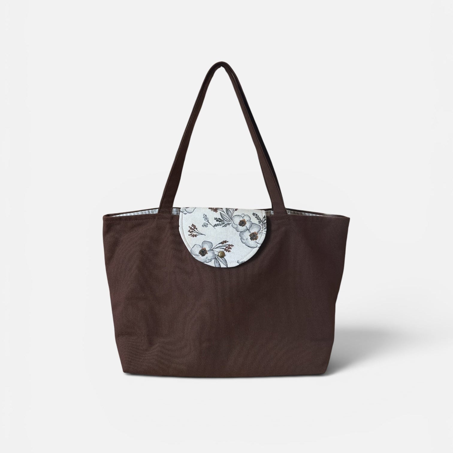 A dark brown tote bag with a circular white floral patch on the front and two shoulder straps, photographed against a clean white background.